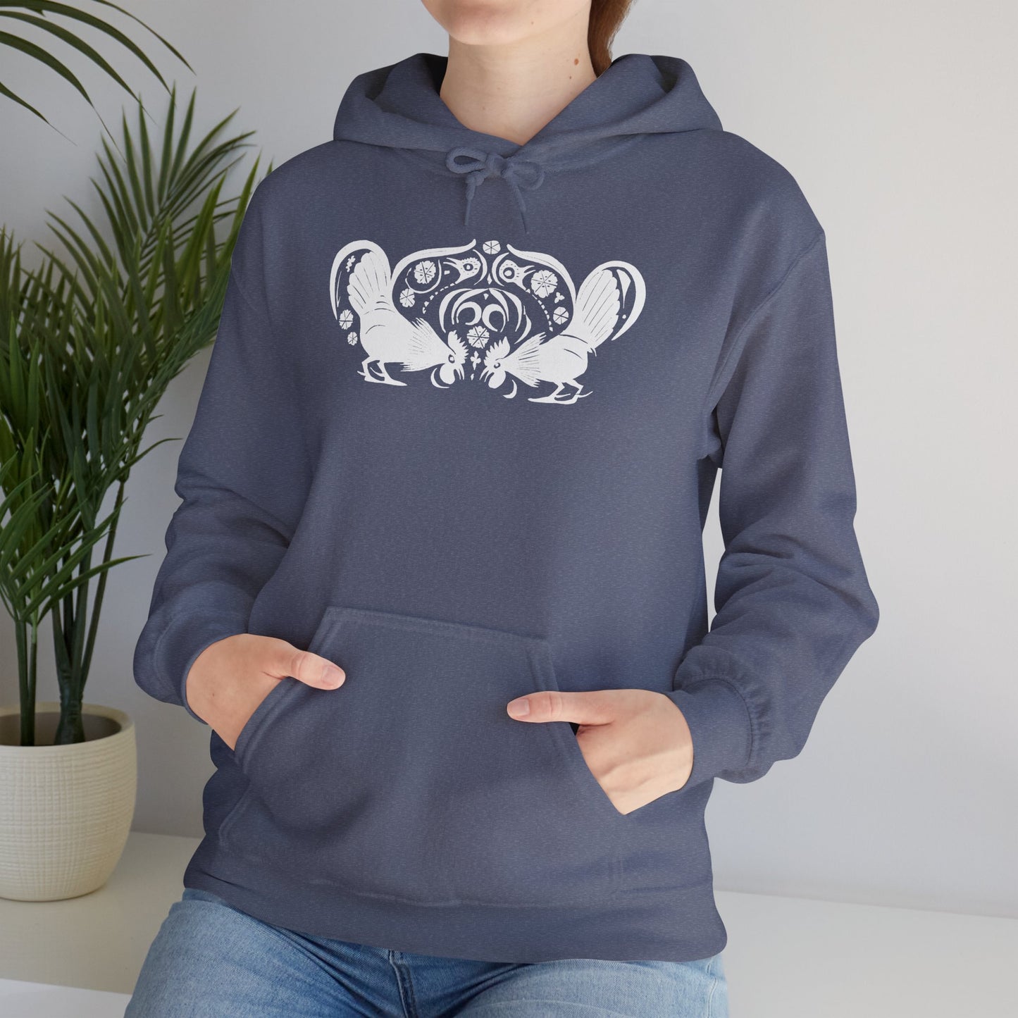 A woman wears a heather navy hoodie with a white, art noveau style print of two roosters crouching face-to-face. The roosters are mirror images of each other and a symmetrical pattern arches between them.