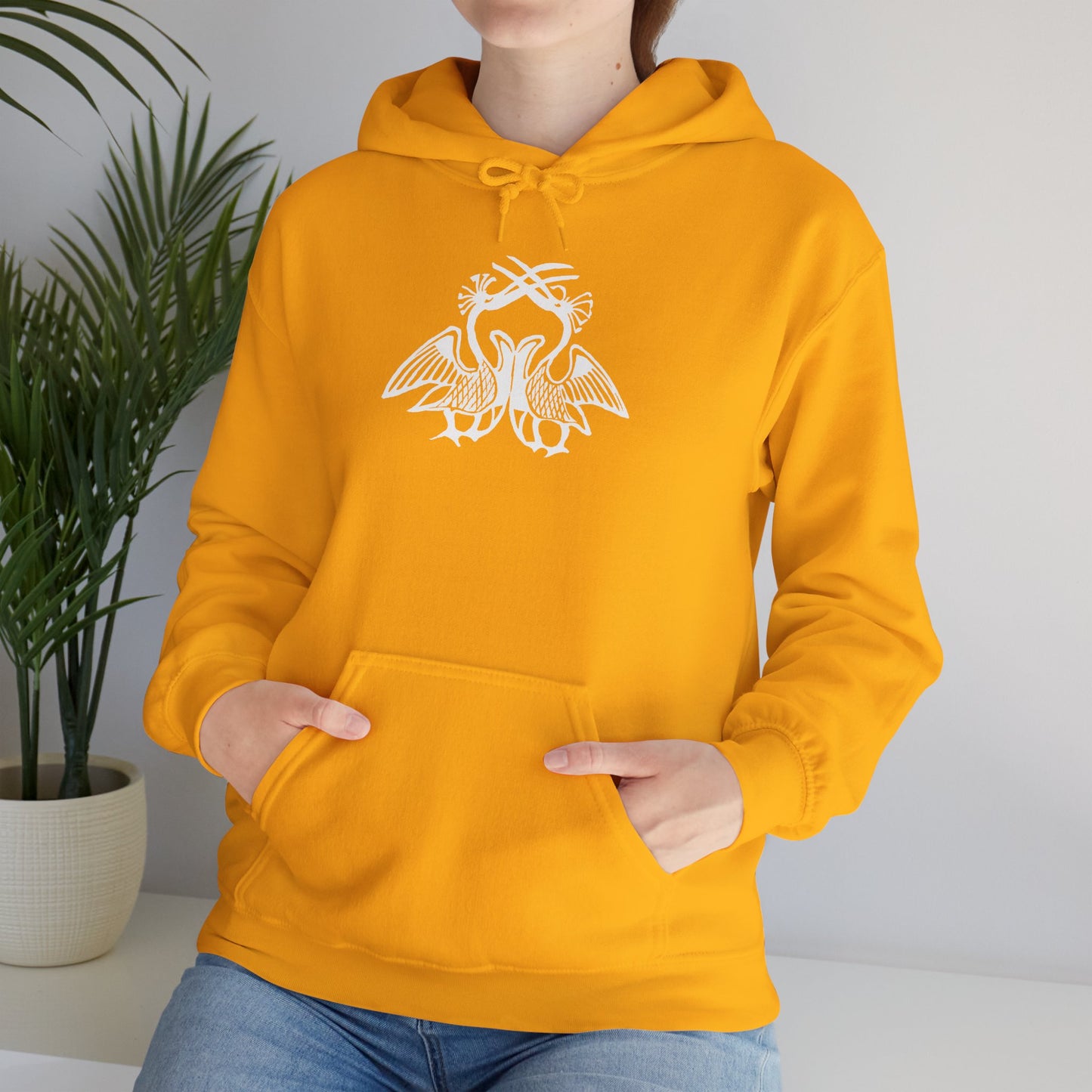A woman wears a deep yellow hoodie with a white, art noveau style print of two birds standing face-to-face. The birds are mirror images of each other and their long beaks cross at the top of the design.