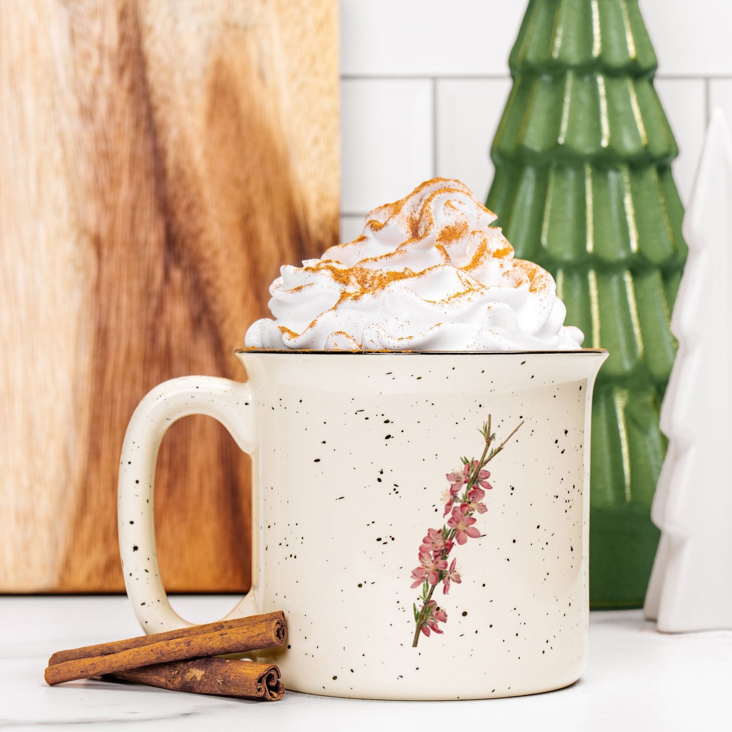 The front view of a cream colored ceramic camp mug with black speckles and a black rim. The mug features a botanical illustration of a branch full of peach blossoms. The mug is filled and topped with whipped cream and a stick of cinnamon sits next to it.