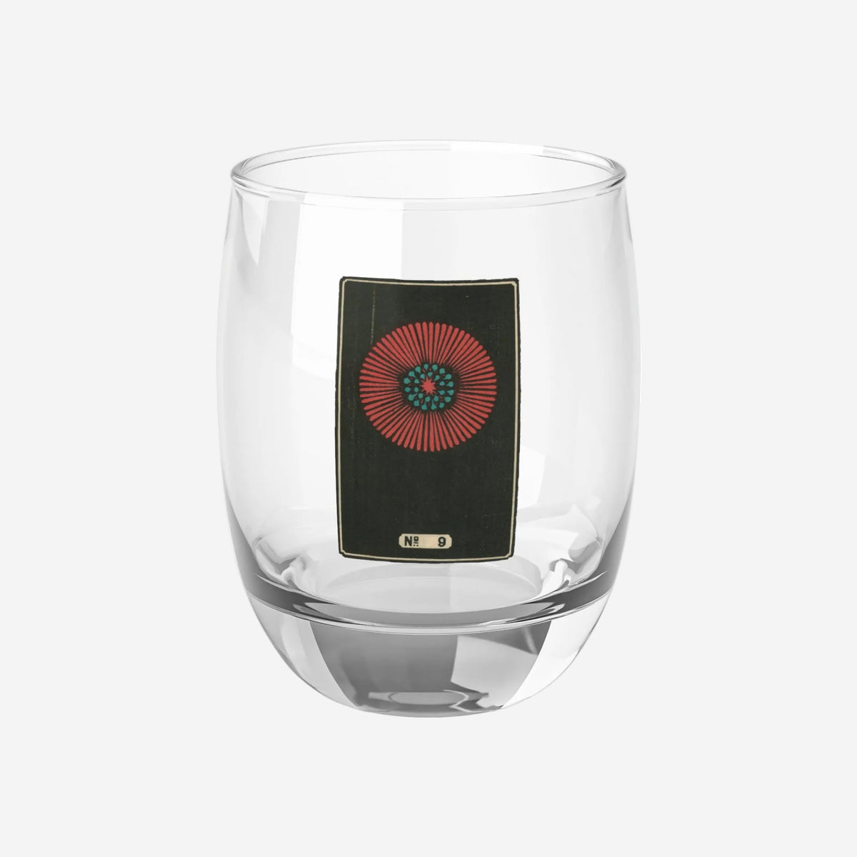 Clear whiskey glass displaying a dark rectangular plate with a crimson and turquoise sunburst woodblock firework print.