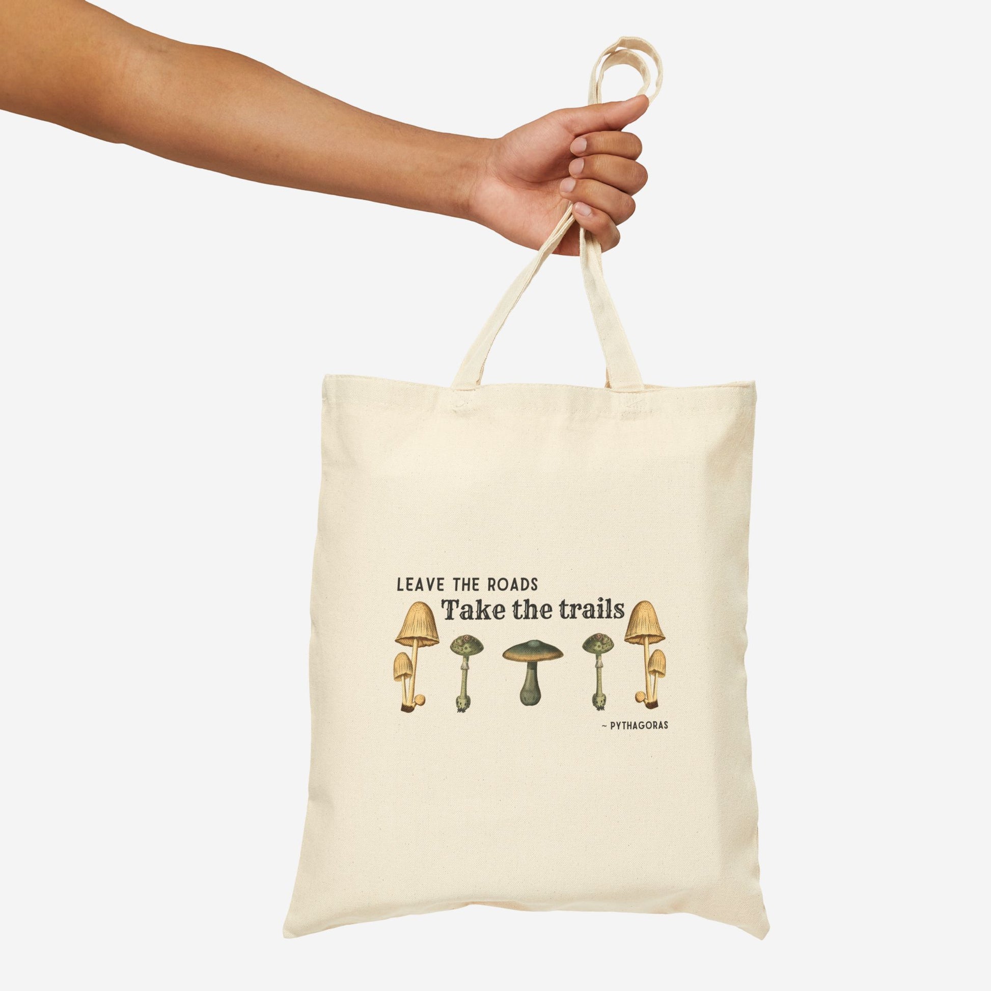 A hand holds a canvas tote bag, the text on the bag reads "Leave the roads take the trails - Pythagoras". There is a symmetrical row of botanical illustrations of mushrooms below the quote.