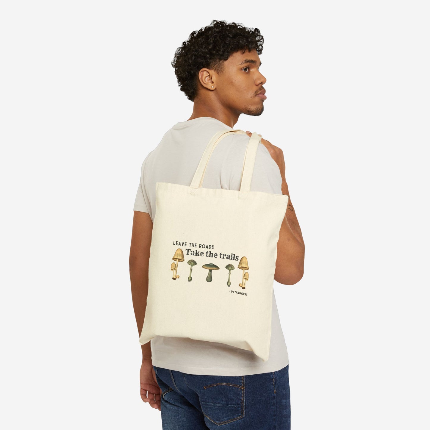 A man holds a natural canvas tote bag over his shoulder, the text on the bag reads "Leave the roads take the trails - Pythagoras". There is a symmetrical row of botanical illustrations of mushrooms below the quote.