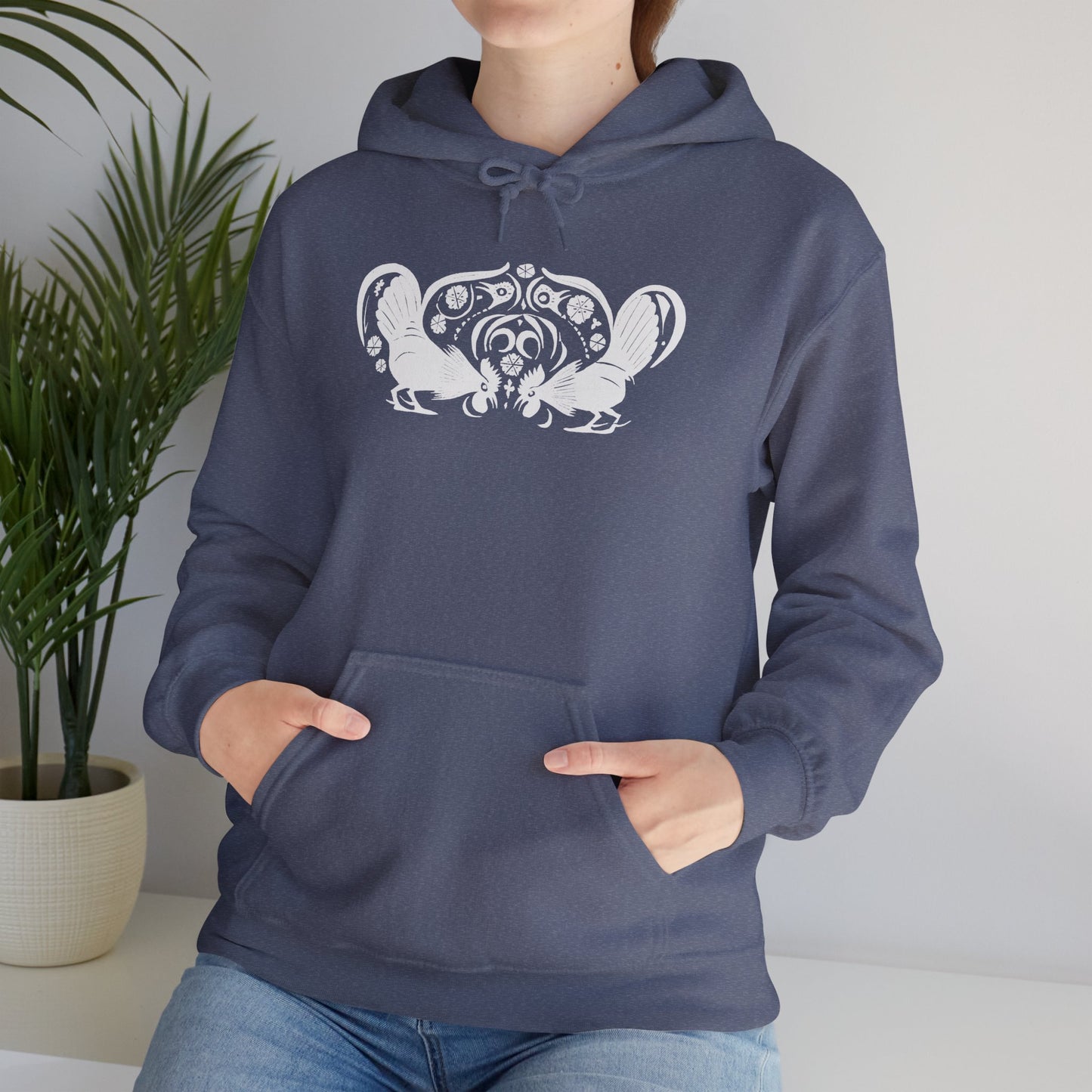 A woman wears a heather navy hoodie with a white, art noveau style print of two roosters crouching face-to-face. The roosters are mirror images of each other and a symmetrical pattern arches between them.