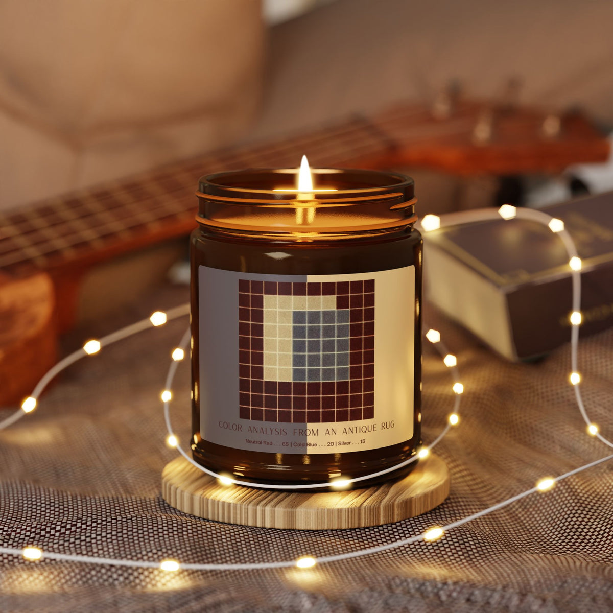 Amber glass candle jar with a black lid and a label showing a ten-by-ten grid of deep red squares surrounding a block of slate blue and a pale cream section, titled Color Analysis from an Antique Rug.