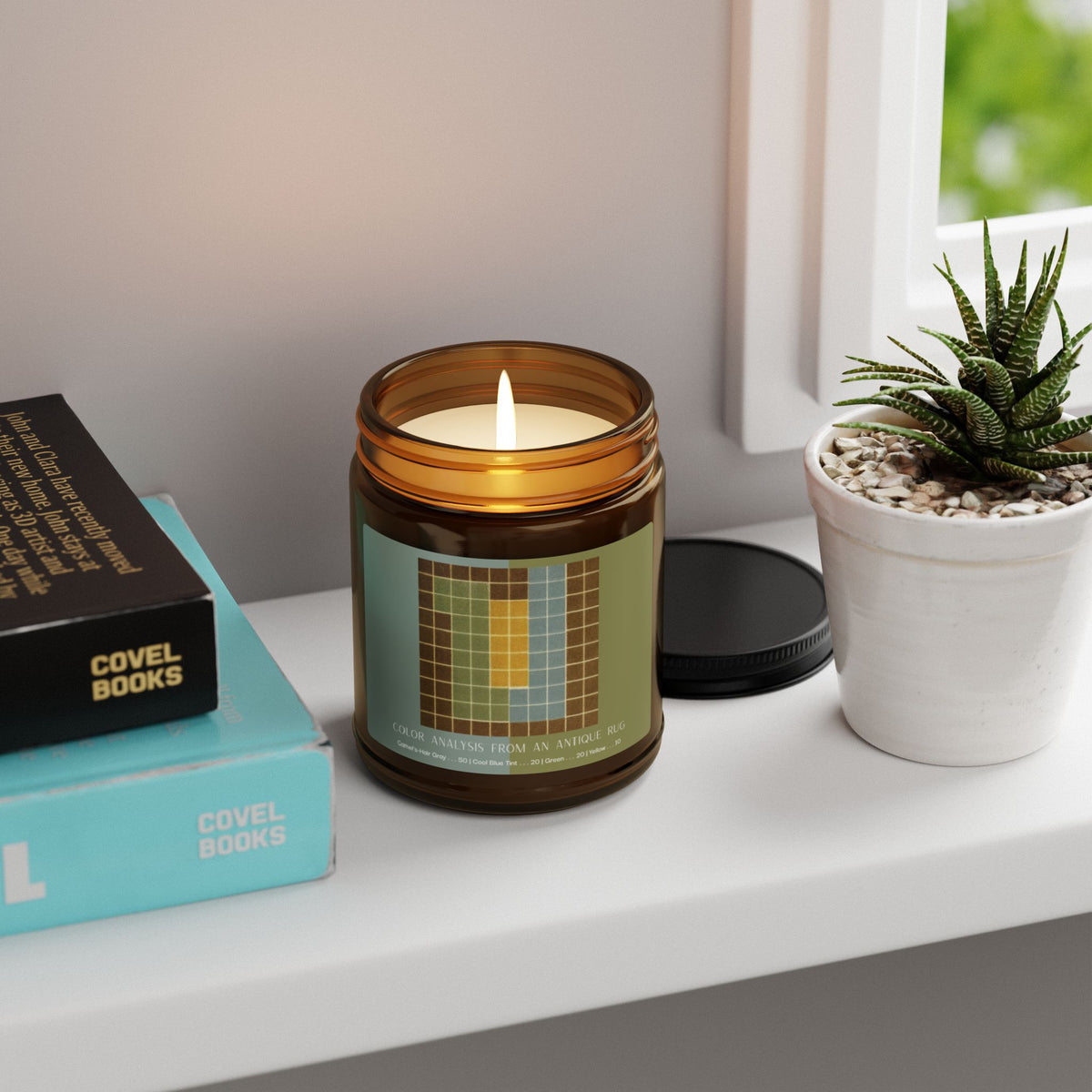 Amber glass candle jar with a black lid and a label showing a ten-by-ten grid with a camel-brown border around columns of sage green, pale blue, and yellow, titled Color Analysis from an Antique Rug.