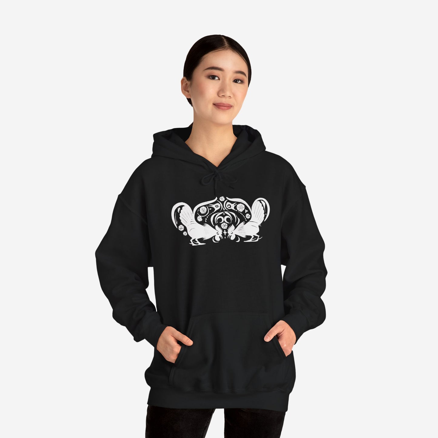 A woman wears a black hoodie with a white, art noveau style print of two roosters crouching face-to-face. The roosters are mirror images of each other and a symmetrical pattern arches between them.