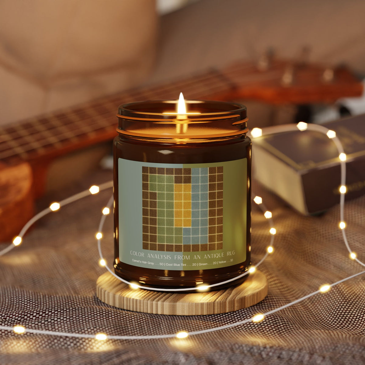 Amber glass candle jar with a black lid and a label showing a ten-by-ten grid with a camel-brown border around columns of sage green, pale blue, and yellow, titled Color Analysis from an Antique Rug.
