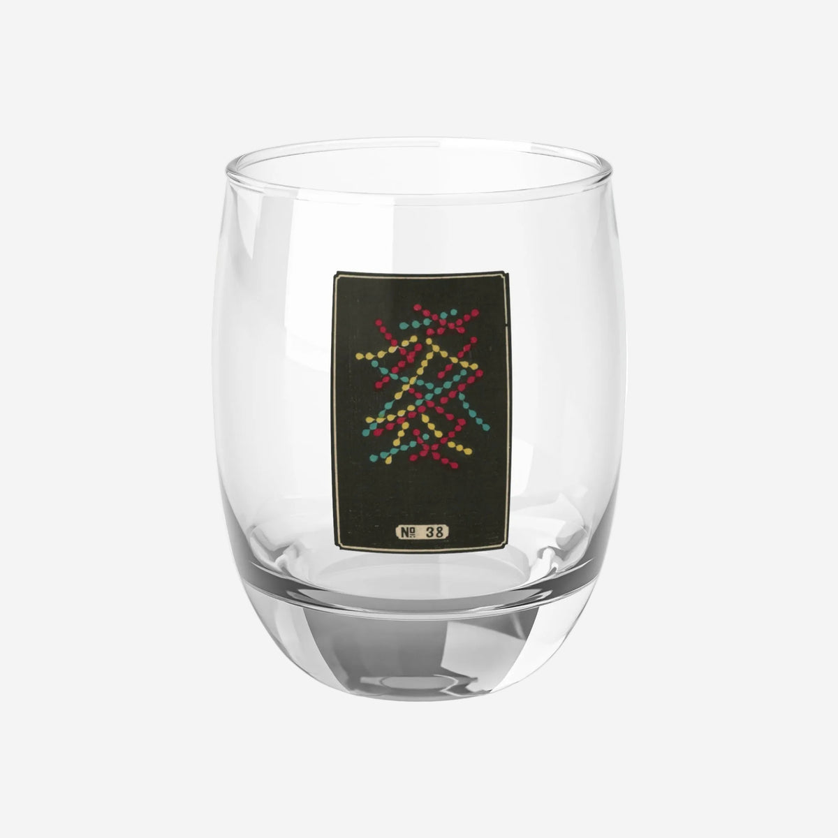 Clear whiskey glass displaying a dark rectangular plate with an intersecting red, yellow, and turquoise lattice woodblock firework print.