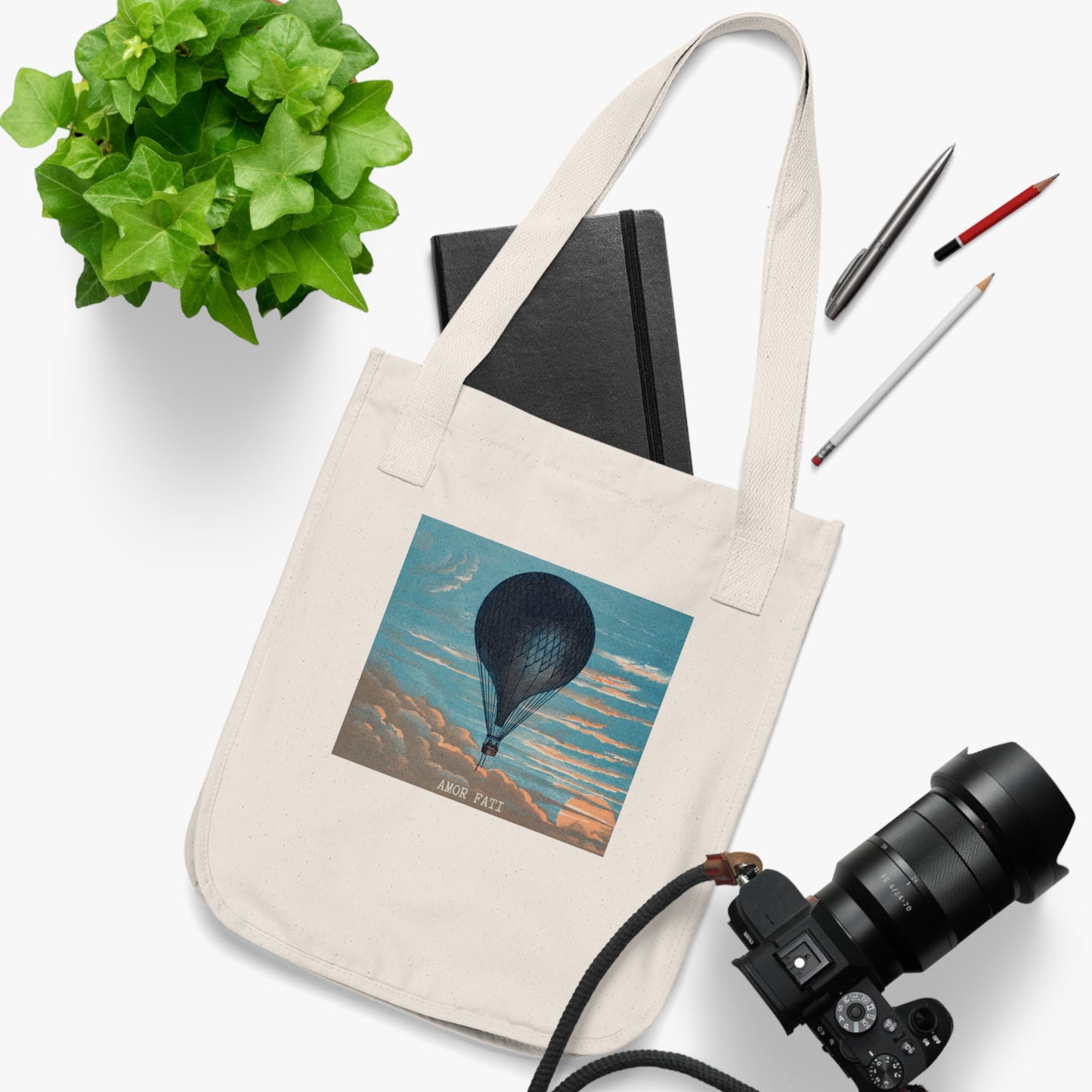 A natural canvas tote bag featuring a hot air balloon floating above peach-toned clouds in a blue sky, with the text "AMOR FATI" beneath. A black notebook, camera, pens, and a green potted plant are arranged around the bag.