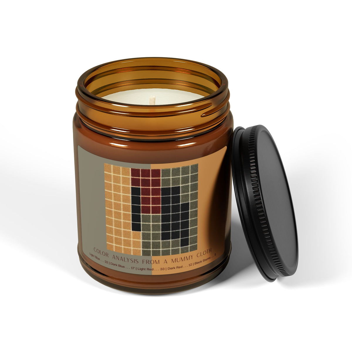 Amber glass candle jar with a black lid and a label showing a ten-by-ten grid of ochre, gray-green, dark red, and black squares, titled Color Analysis from a Mummy Cloth.