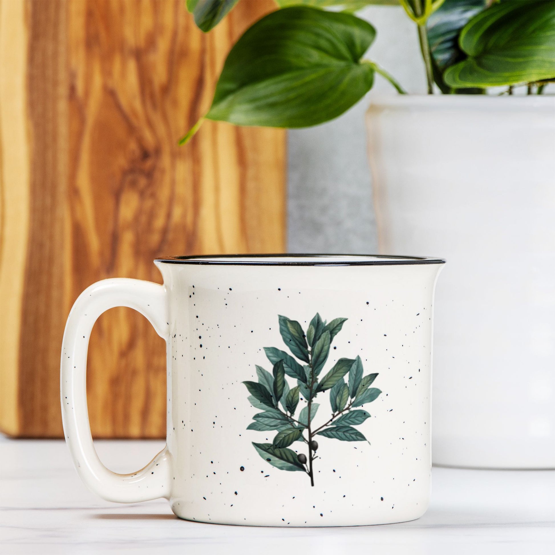 The front view of a cream colored ceramic camp mug with black speckles and a black rim. The mug features a botanical illustration of a branch of bay leaves.