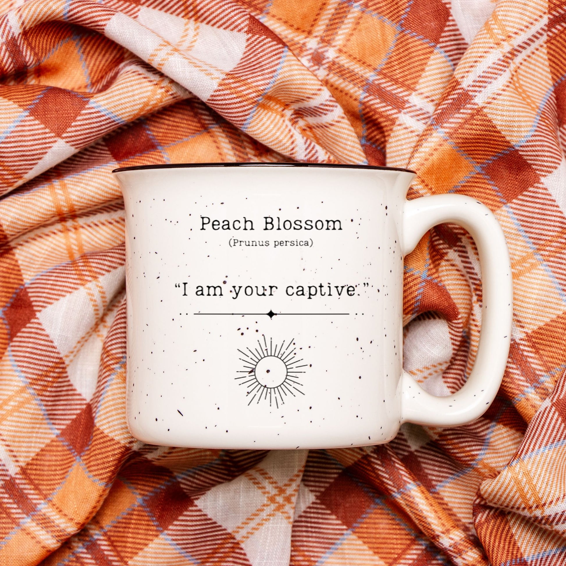 The back view of a cream colored ceramic camp mug with black speckles and a black rim. Text on the mug reads Peach Blossom (Prunus persica) "I am your captive". The text is printed above a black outline of a sun.