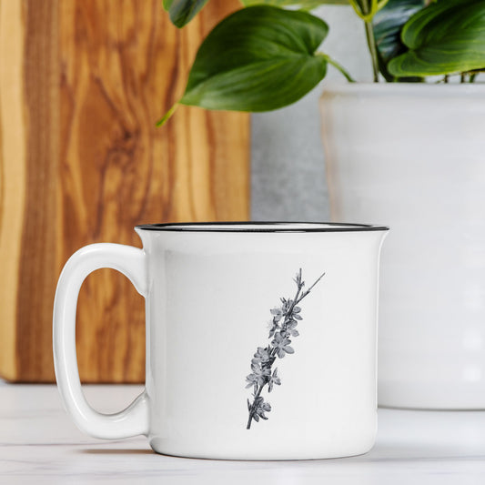 The front view of a white ceramic camp mug with a black rim. The mug features a black and white botanical illustration of a branch full of peach blossoms.