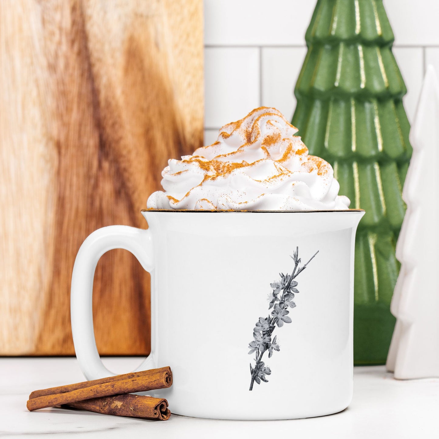 The front view of a white ceramic camp mug with a black rim. The mug features a black and white botanical illustration of a branch full of peach blossoms. The mug is filled and topped with whipped cream and a stick of cinnamon sits next to it.