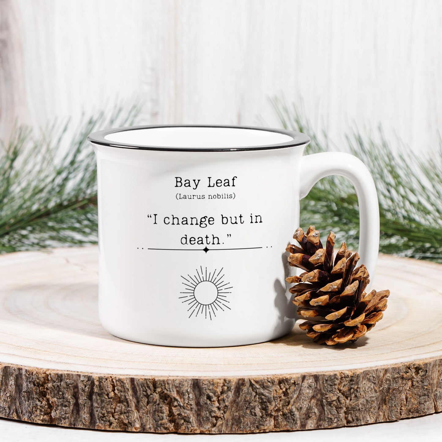 The back view of a white ceramic camp mug with a black rim. Text on the mug reads Bay Leaf (Laurus nobilis) "I change but in death." The text is printed over a black outline of a sun. The mug sits on a wood slab with a pinecone leaned against it.