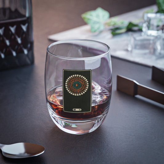 Clear whiskey glass displaying a dark rectangular plate with a cream, orange, and turquoise woodblock firework print.