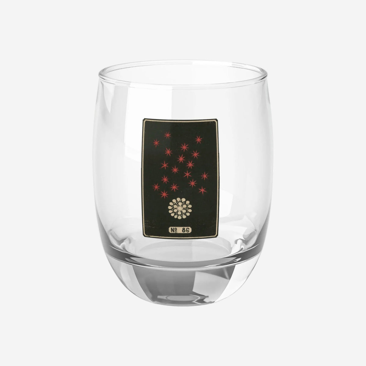 Clear whiskey glass displaying a dark rectangular plate with an ivory burst and rising red stars woodblock firework print.