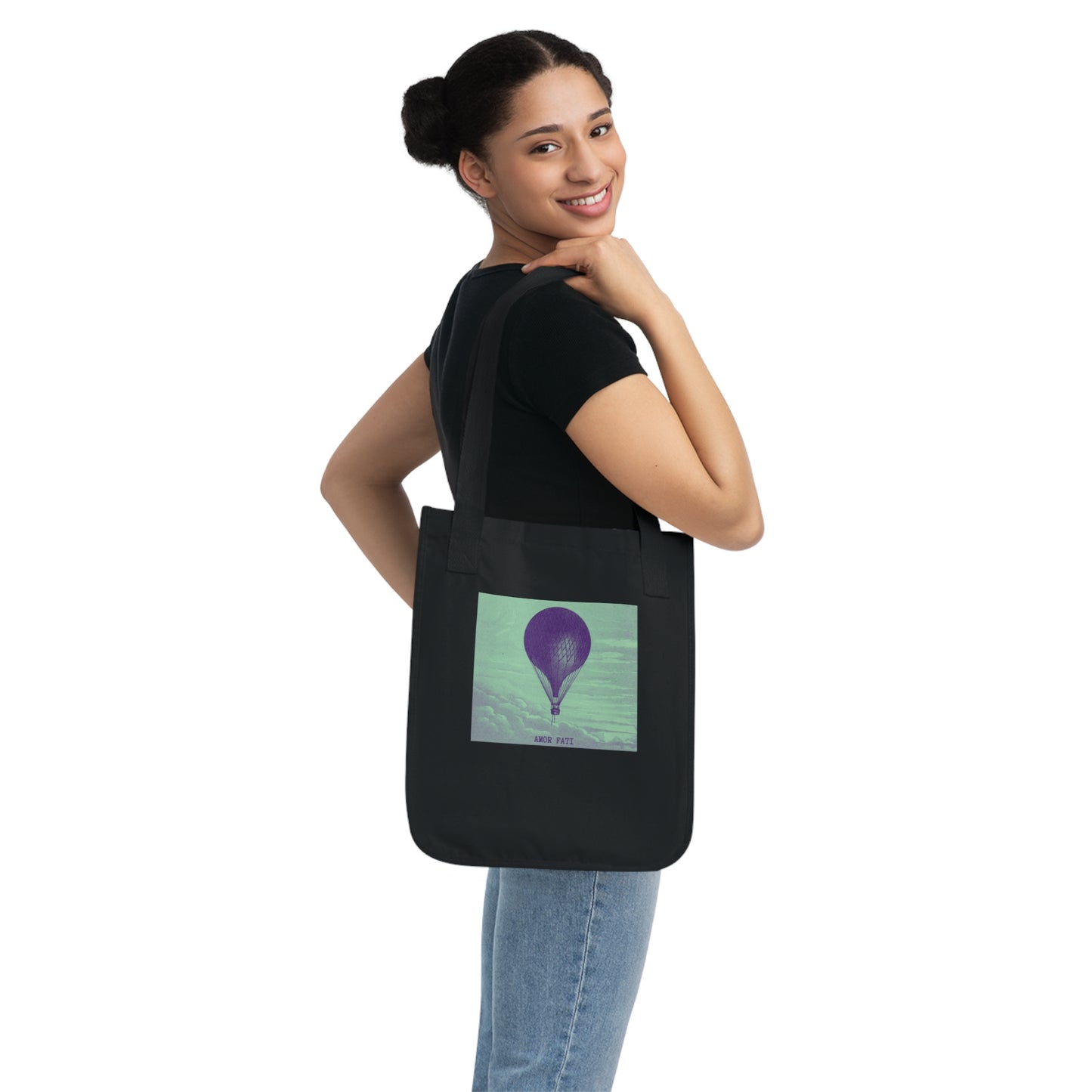 A woman wearing a black t-shirt and jeans carries a black canvas tote bag over her shoulder. The bag features a purple hot air balloon against a mint green sky, with the text "AMOR FATI" beneath.