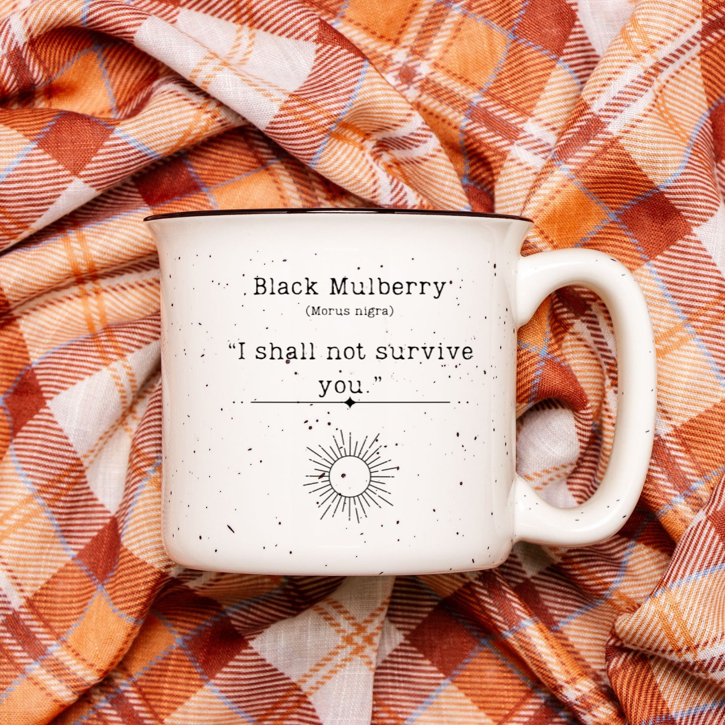 The back view of a cream colored ceramic camp mug with black speckles and a black rim. Text on the mug reads Black Mulberry (Morus nigra) "I shall not survive you". The text is printed above a black outline of a sun.