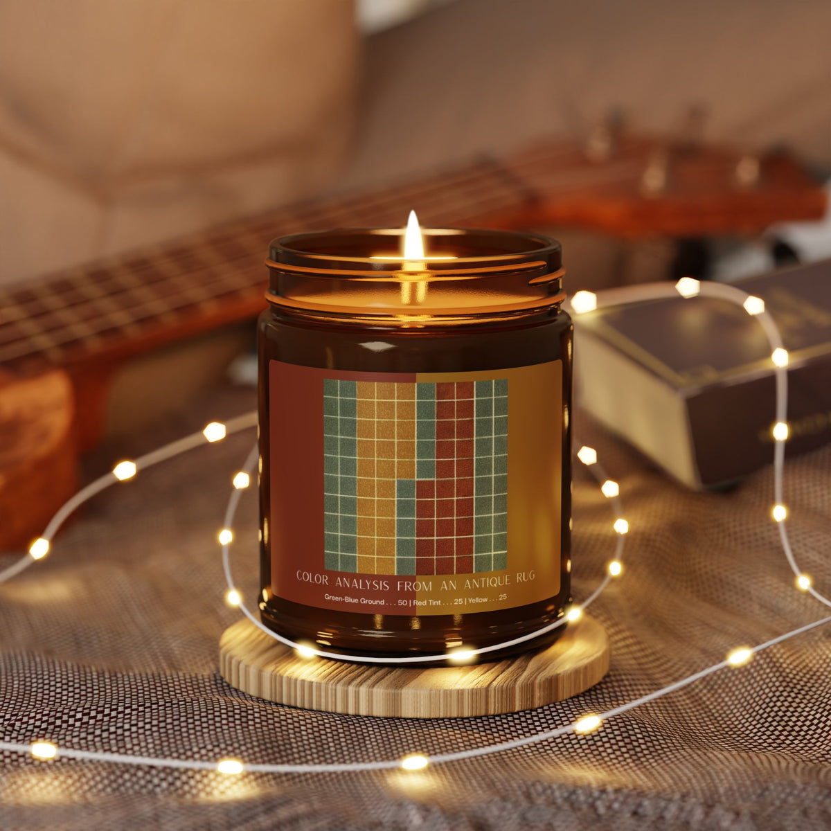 Amber glass candle jar with a black lid and a label showing a ten-by-ten grid of gray-green squares with vertical bands of golden yellow and brick red, titled Color Analysis from an Antique Rug.