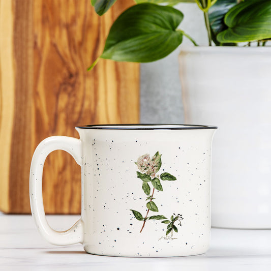 The front view of a cream colored ceramic camp mug with black speckles and a black rim. The mug features a botanical illustration of a Laurestina flower.