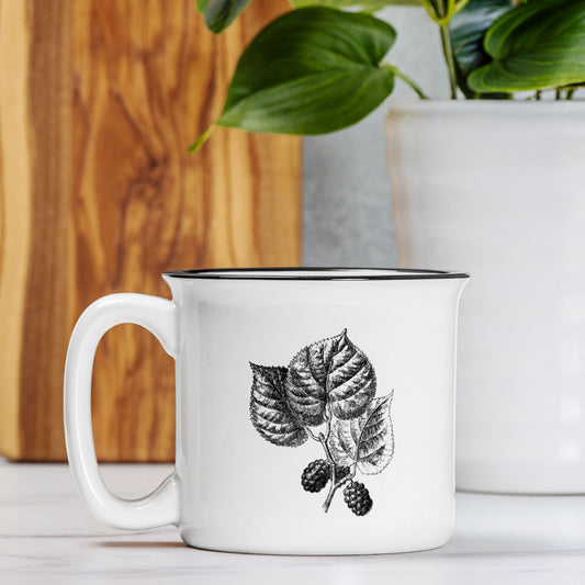 The front view of a white ceramic camp mug with a black rim. The mug features a black and white botanical illustration of a black mulberry branch.