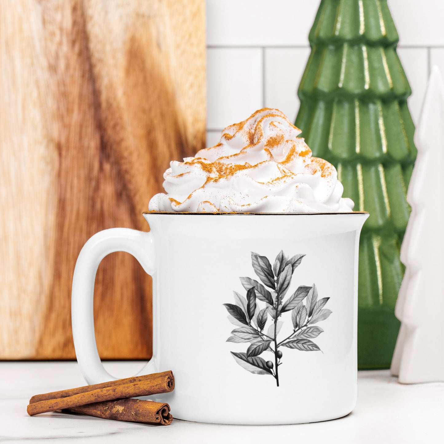 The front view of a white ceramic camp mug with a black rim. The mug features a black and white botanical illustration of a branch of bay leaves. The mug is filled and topped with whipped cream and a stick of cinnamon sits next to it.