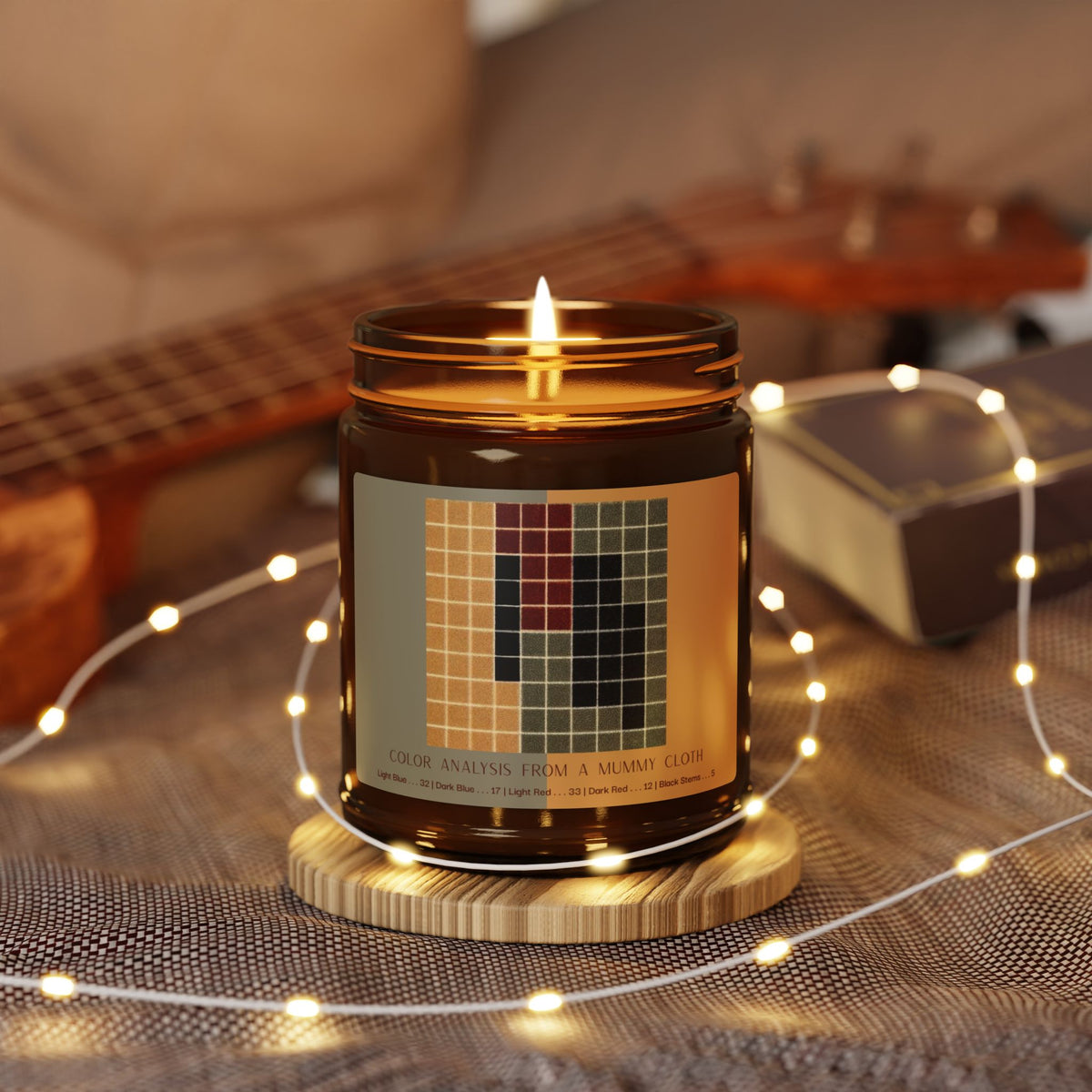 Amber glass candle jar with a black lid and a label showing a ten-by-ten grid of ochre, gray-green, dark red, and black squares, titled Color Analysis from a Mummy Cloth.