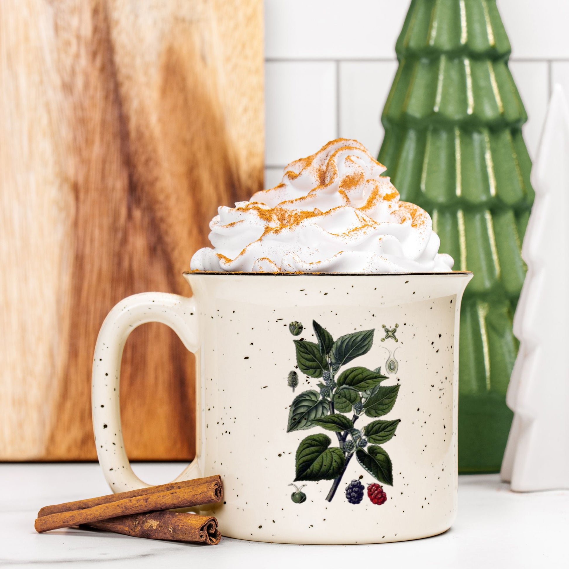 The front view of a cream colored ceramic camp mug with black speckles and a black rim. The mug features a botanical illustration of a black mulberry branch. The mug is filled and topped with whipped cream and a stick of cinnamon sits next to it.