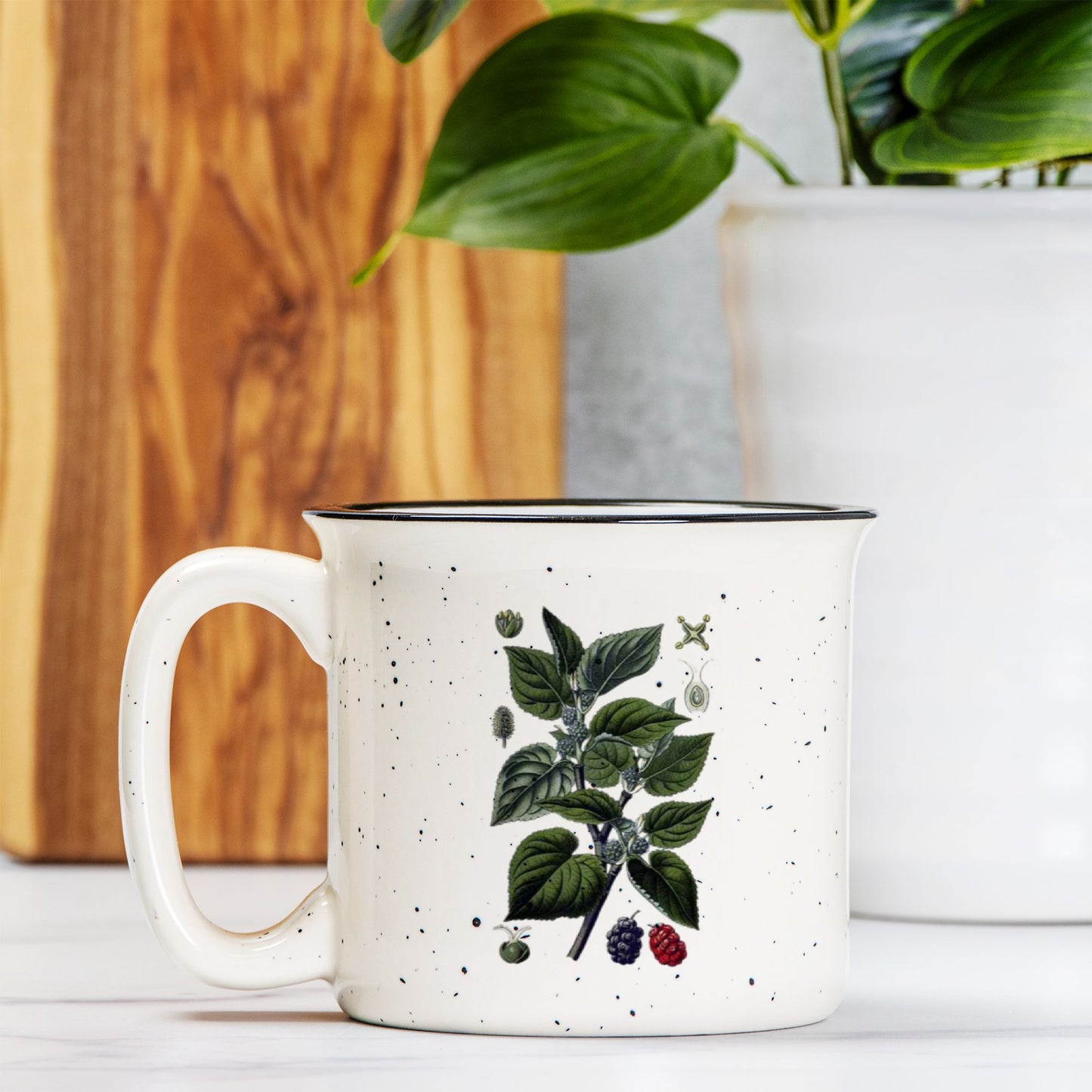 The front view of a cream colored ceramic camp mug with black speckles and a black rim. The front of the mug features a botanical illustration of a black mulberry branch.