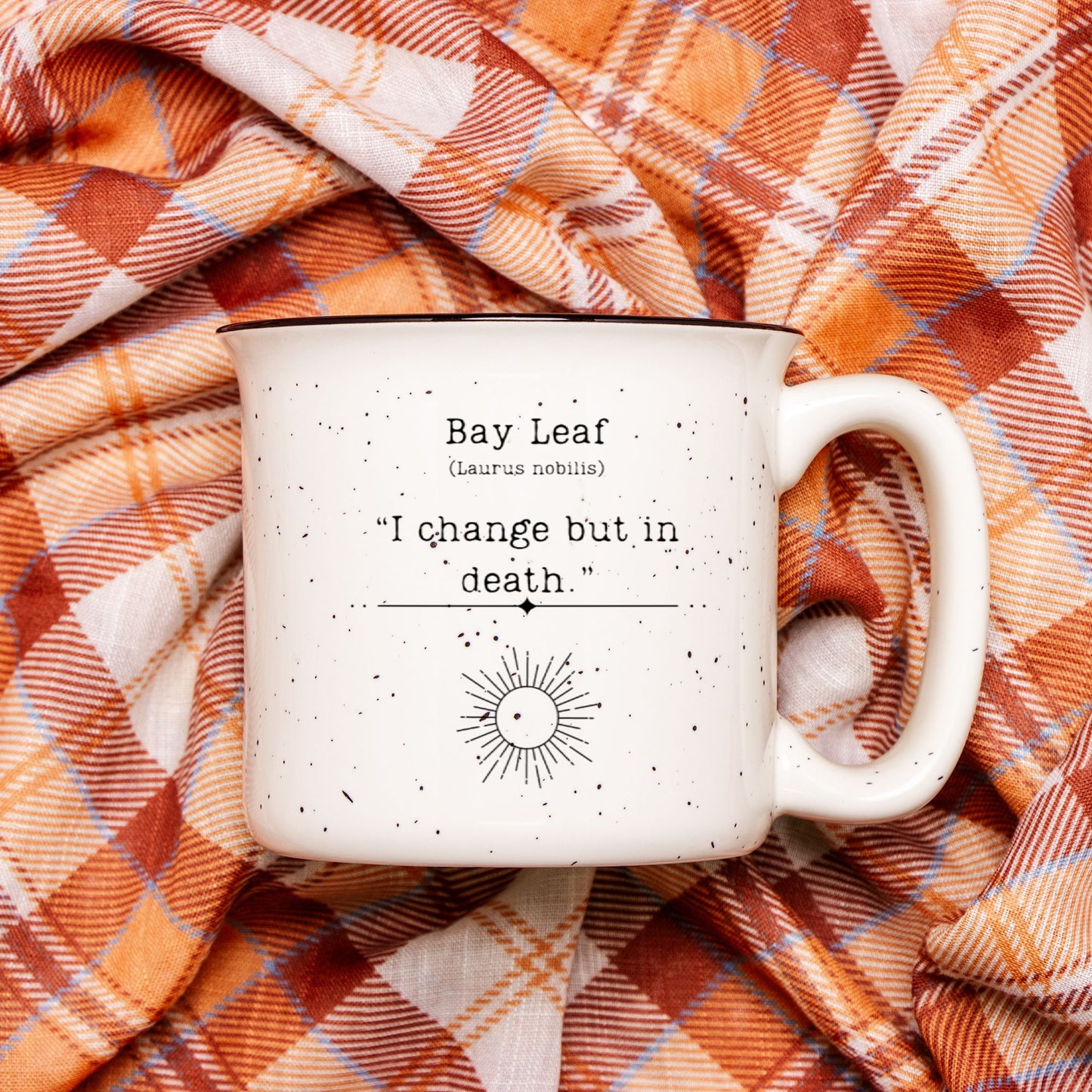 The back view of a cream colored ceramic camp mug with black speckles and a black rim. Text on the mug reads Bay Leaf (Laurus nobilis) "I change but in death." The text is printed over a black outline of a sun. 