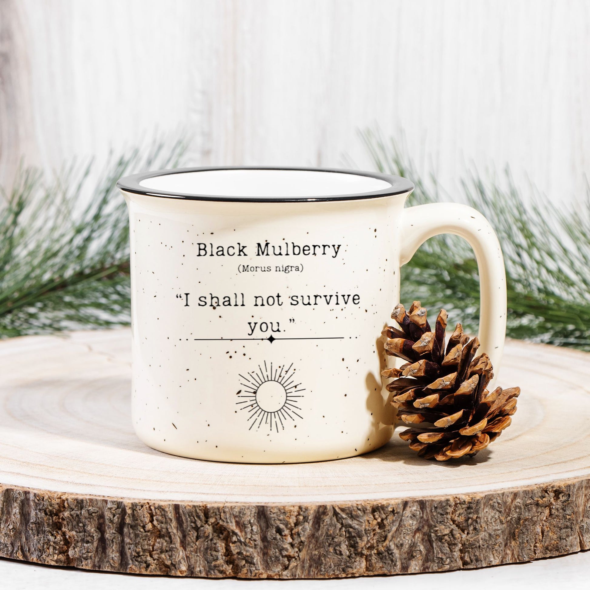 The back view of a cream colored ceramic camp mug with black speckles and a black rim. Text on the mug reads Black Mulberry (Morus nigra) "I shall not survive you". The text is printed above a black outline of a sun. The mug sits on a wood slab with a pinecone leaning against it.