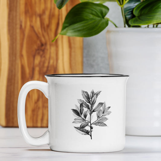 The front view of a white ceramic camp mug with a black rim. The mug features a black and white botanical illustration of a branch of bay leaves.