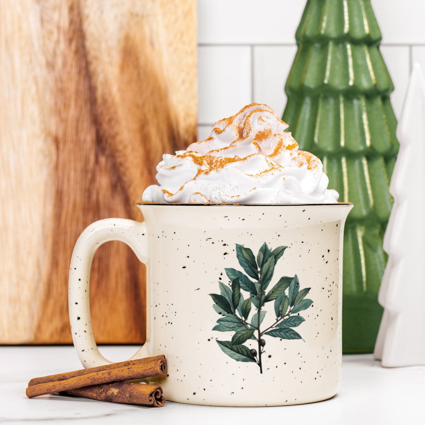 A cream colored ceramic camp mug with a black rim. The mug features a colored botanical illustration of a branch of bay leaves. The mug is filled and topped with whipped cream and a stick of cinnamon sits next to it.