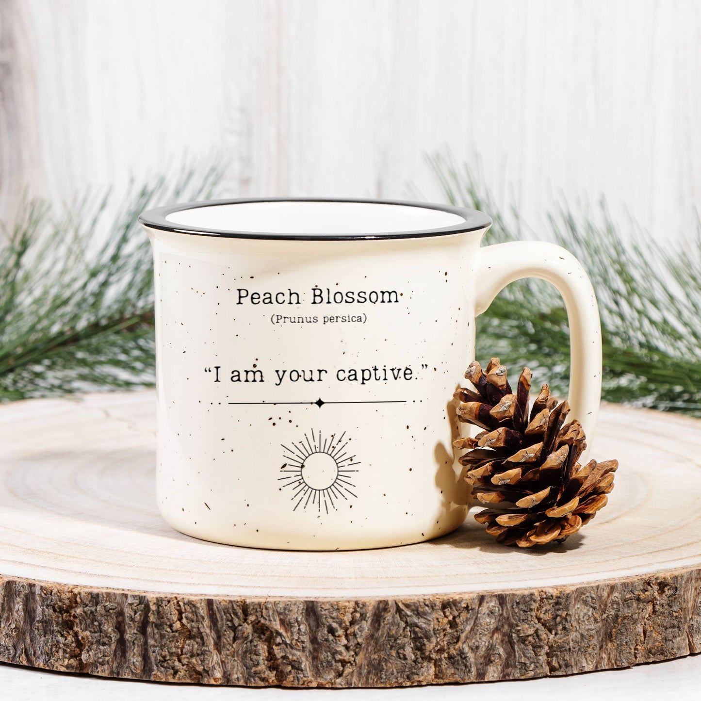 The back view of a cream colored ceramic camp mug with black speckles and a black rim. Text on the mug reads Peach Blossom (Prunus persica) "I am your captive". The text is printed above a black outline of a sun. The mug sits on a wood slab with a pinecone leaning against it.